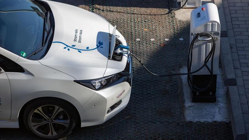 A white electric Nissan Leaf could be seen at the charging at the electric charging station. Bonnet of the car seems to have a sticker of a plug with leaves grown on its cable denoting that its nature/environment friendly alongside a written sticker (strom rein strom raus). Presenting How EVs help in tackling climate change